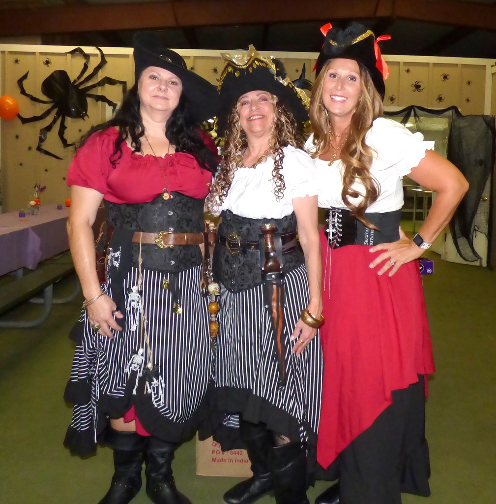 Three women dressed as pirates, wearing black hats, striped skirts, and red or white tops, stand together smiling at a Halloween themed event with spider decorations and cobwebs in the background Three women dressed as pirates, wearing black hats, striped skirts, and red or white tops, stand together smiling at a Halloween themed event with spider decorations and cobwebs in the background