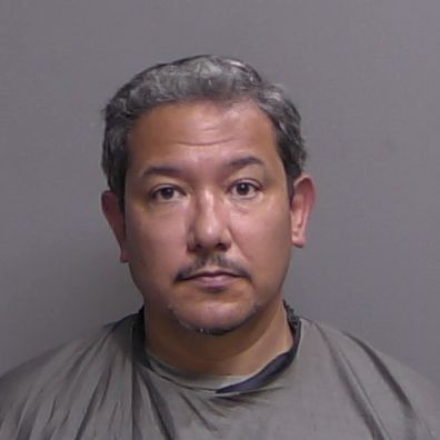 A middle aged man with short, gray black hair and a mustache is facing the camera against a plain gray background, wearing a gray garment that covers his shoulders and upper chest