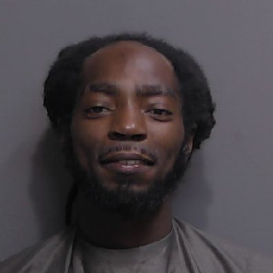 A man with short dreadlocks and a beard is smiling slightly while wearing a gray drape in front of a plain gray background