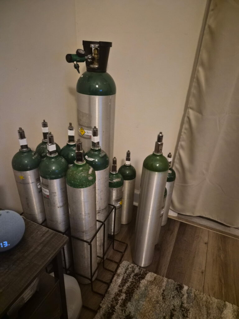 Several green and silver oxygen tanks are standing upright in a corner, some in a metal rack, on a wood floor near a beige rug, a side table, and a curtain