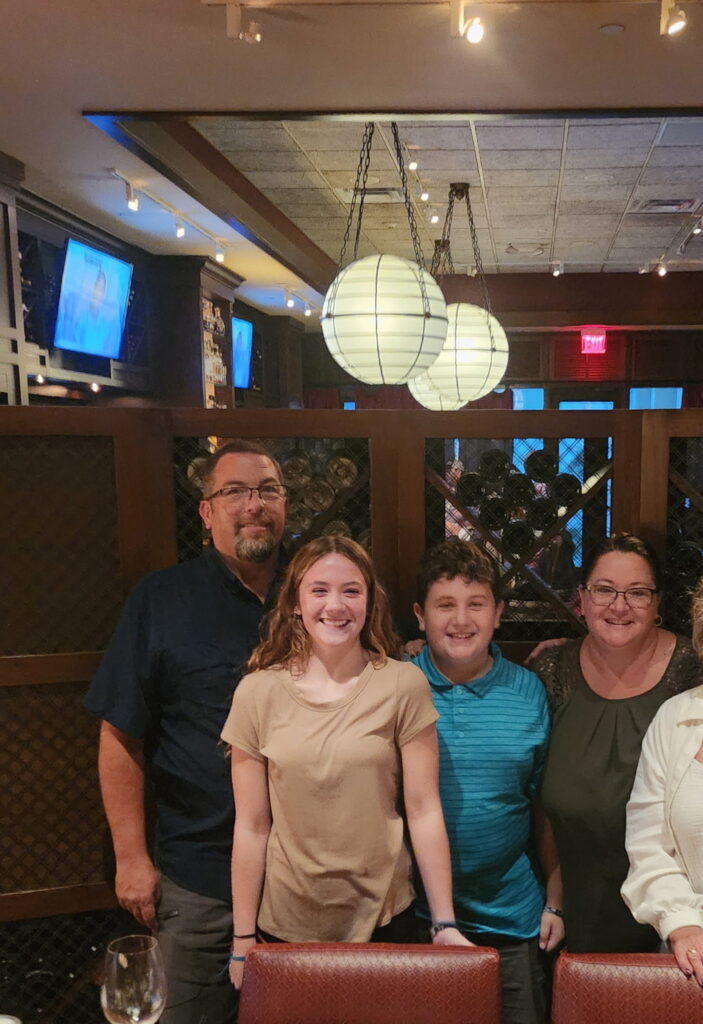 Four people, two adults and two children, stand together inside a restaurant, smiling at the camera Warm lighting and hanging round lamps create a cozy atmosphere A TV and wooden decor are visible in the background