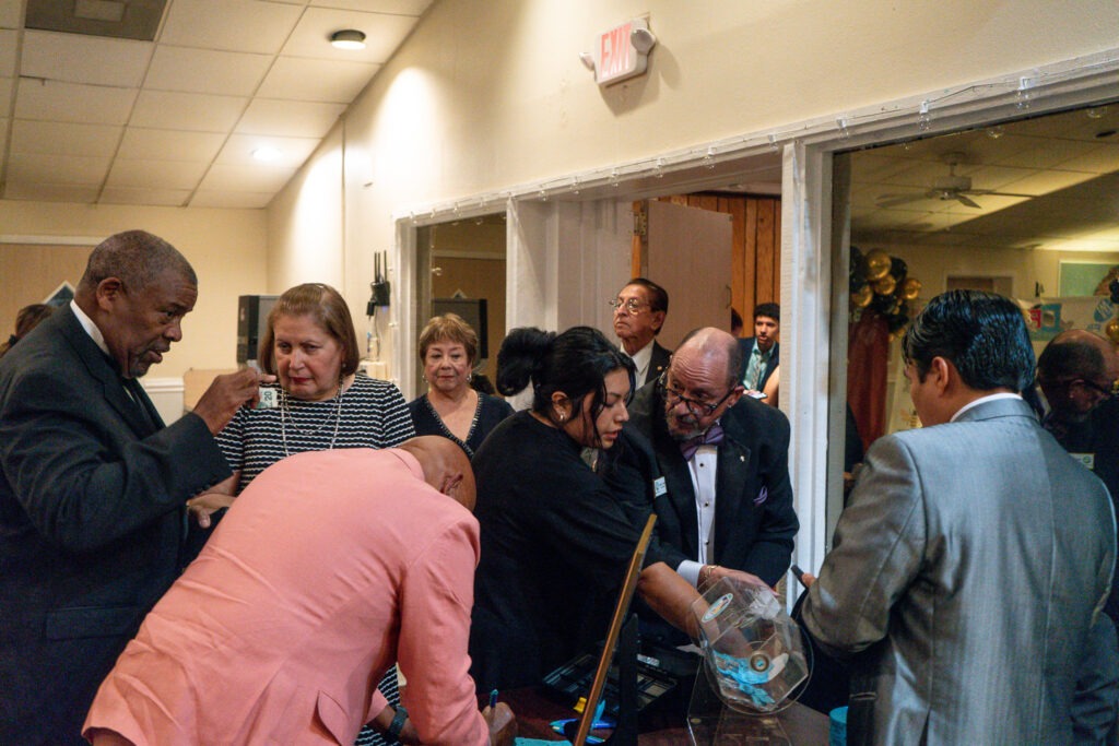 A group of formally dressed people gather around a table at an indoor event, engaging in conversation and examining raffle tickets or papers The atmosphere appears lively and social