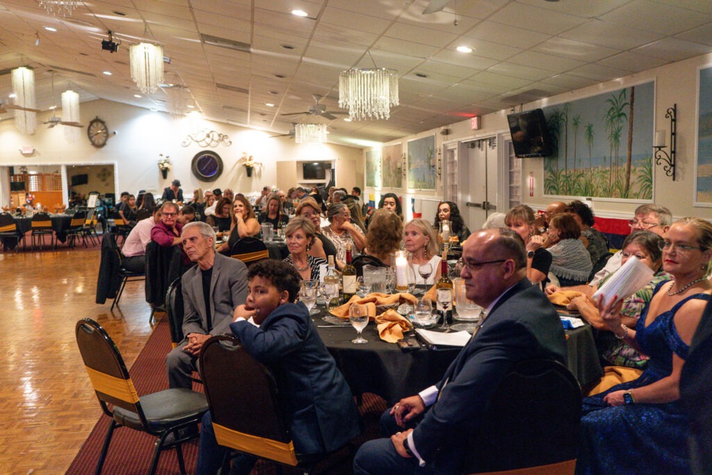 A group of people dressed in formal attire sit around tables at a banquet hall, attentively watching something off camera The room is decorated with chandeliers, palm tree murals, and white walls
