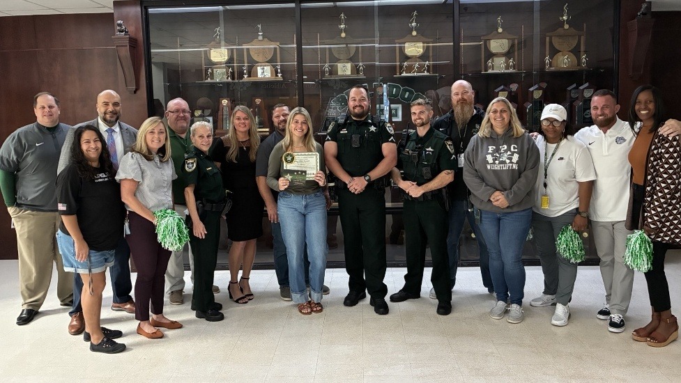 A group of adults, including uniformed officers and civilians, pose smiling indoors in front of a trophy case One woman in the center holds a certificate, and two people hold green and white pom poms