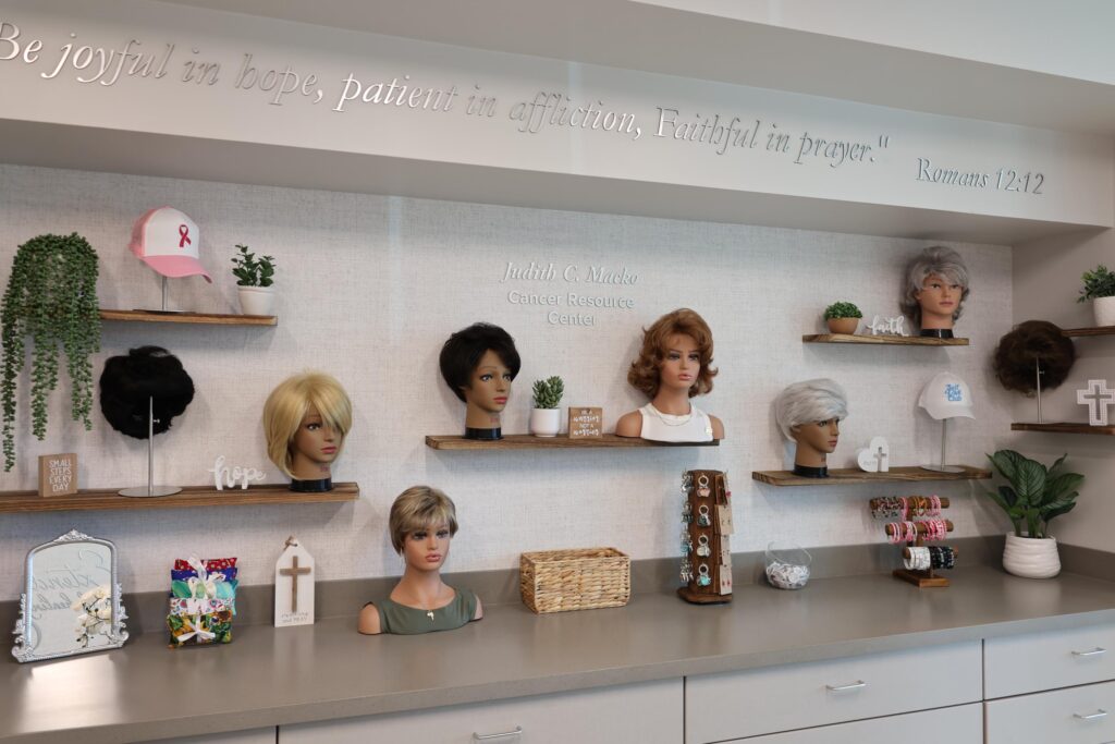 A display of wigs on mannequin heads is arranged on shelves in a resource center, along with hats, inspirational plaques, and support bracelets A Bible verse is inscribed on the wall above the shelves