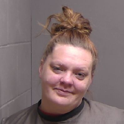 A woman with light skin and brown hair in a messy bun wears a dark shirt and stands against a gray wall, looking at the camera with a slight smile