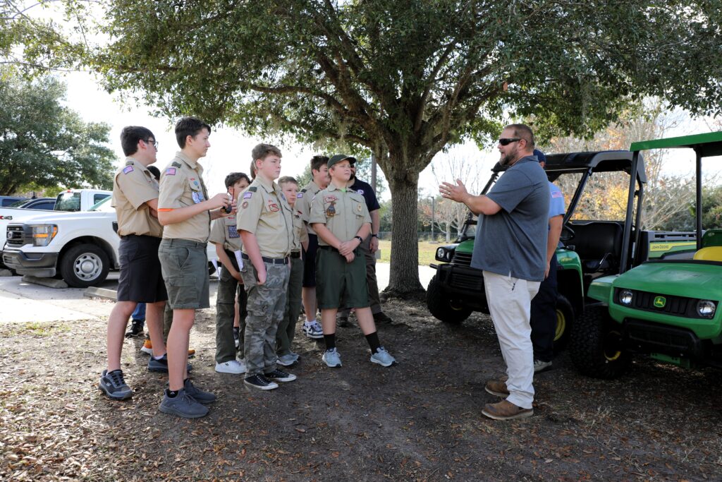 A group of Boy Scouts in uniform stand outdoors near trees and utility vehicles, listening attentively to an adult man who is speaking to them