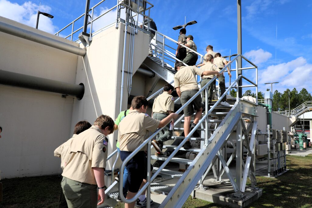 A group of scouts in uniform climb an outdoor metal staircase at an industrial facility on a sunny day, with blue sky and scattered clouds in the background