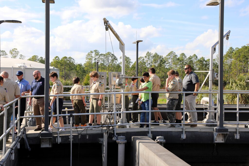 A group of people, including adults and youths in uniforms, stand on a raised platform at an outdoor industrial facility with metal railings, machinery, and trees in the background