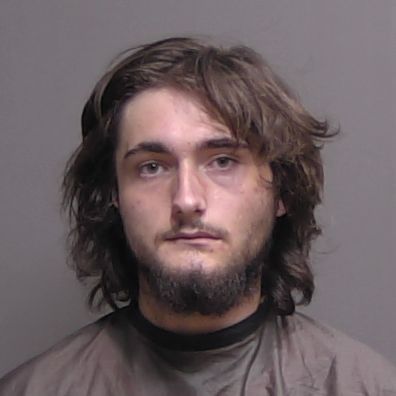 A young man with long brown hair and a partial beard looks at the camera with a neutral expression, wearing a dark cape against a plain gray background