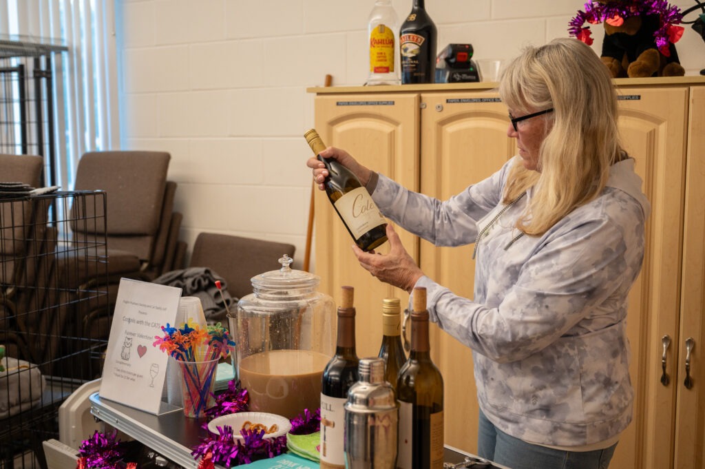 A woman with long blonde hair and glasses holds a bottle of white wine, preparing to pour it She stands by a table with more wine bottles, a drink dispenser, and festive decorations in a room with cabinets and chairs