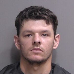 A young man with short dark hair and light skin looks directly at the camera with a neutral expression, wearing a black garment against a plain gray background