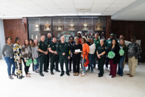 A group of people, including police officers in uniform and civilians, pose together indoors in front of a trophy case Some hold green pom poms, and one person is holding a cake box
