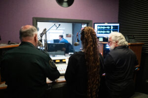 Three people sit at a radio station control panel with microphones, facing a window showing another person at a desk in a separate studio Audio waveforms are displayed on a monitor The room has purple walls and soundproofing panels