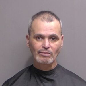A middle aged man with short hair, slight facial hair, and a neutral expression faces the camera against a plain background, wearing a dark garment typically used for mugshots
