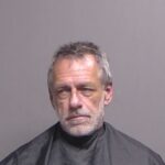 A middle aged man with short gray hair and a beard, wearing a black smock, is photographed against a plain gray background