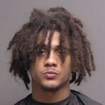 A young man with medium brown skin and shoulder length, curly dreadlocks stares straight ahead with a neutral expression He is wearing a dark colored top and poses against a plain gray background
