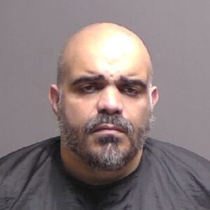 A man with a shaved head and beard wearing a black drape looks directly at the camera against a plain gray background
