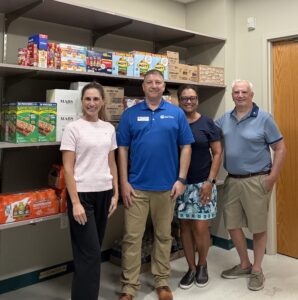 Four adults stand smiling in front of shelves stocked with snacks and drinks in a pantry or storage room The group is casually dressed and standing on a tile floor near a wooden door Four adults stand smiling in front of shelves stocked with snacks and drinks in a pantry or storage room The group is casually dressed and standing on a tile floor near a wooden door