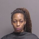 A woman with long, braided hair styled in a high ponytail poses against a plain background, wearing a black cape commonly used for mugshots She has a neutral facial expression