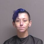 A person with short, partially blue hair, wearing a dark top, is photographed against a plain background They have a neutral facial expression A person with short, partially blue hair, wearing a dark top, is photographed against a plain background They have a neutral facial expression