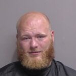 A man with a reddish blond beard and shaved head, wearing a dark cape or smock, stands against a plain gray background, facing the camera A man with a reddish blond beard and shaved head, wearing a dark cape or smock, stands against a plain gray background, facing the camera