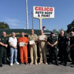 Nine people stand in front of an old sheriff’s vehicle and a Celico Auto Body & Paint sign Three people in the middle wear prison uniforms and hold certificates, while others are in uniforms or plain clothes