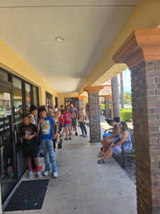 A group of children and adults stand in a line outside a building under a covered walkway, while some kids sit on a bench nearby Bright daylight and greenery are visible in the background