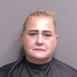 A woman with blonde hair pulled back, wearing a black garment, is photographed against a plain gray background She has a neutral expression and is facing the camera directly