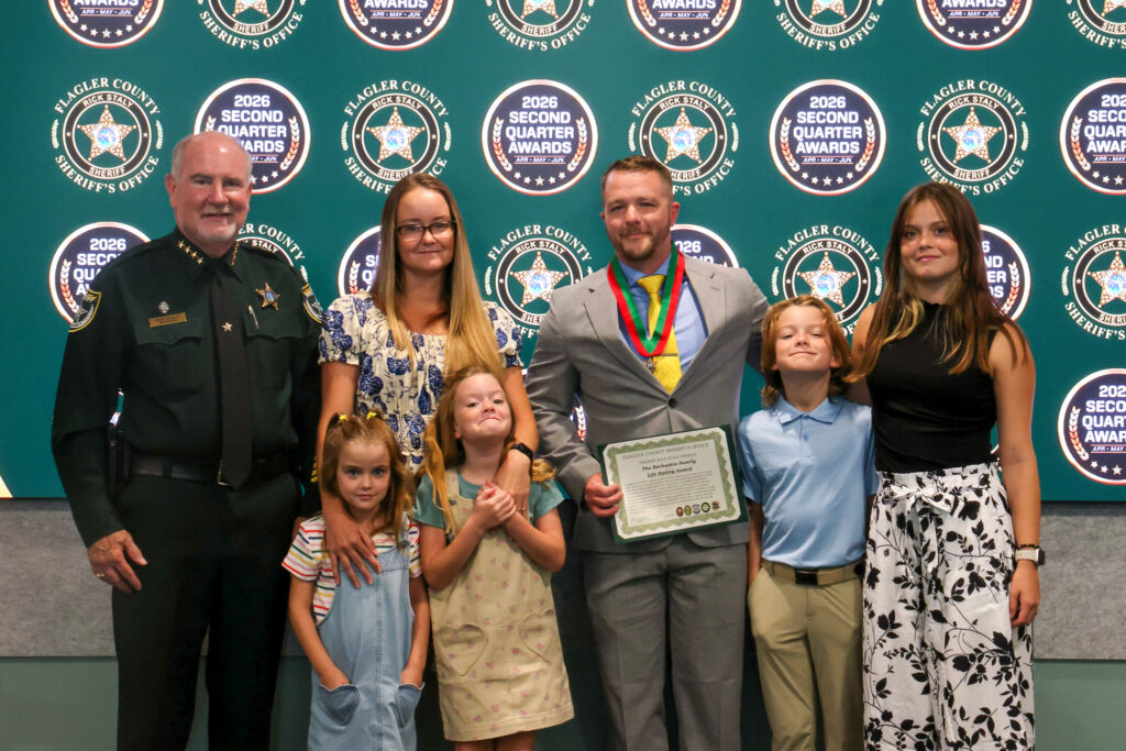 A group of seven people, including a uniformed officer, pose indoors at a Flagler County awards event A man holds a certificate and wears medals, surrounded by four children and two women, with award signs in the background