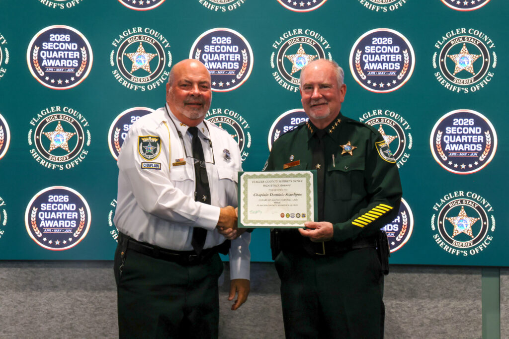 Two uniformed officers shake hands while one presents a certificate They stand in front of a backdrop featuring sheriff badges and logos for the Flagler County Sheriff’s Office Second Quarter Awards