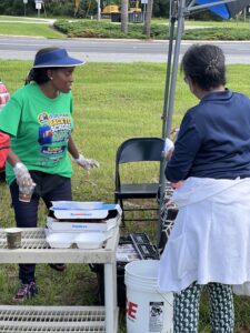 Two women stand near a table with pizza boxes and drinks at an outdoor event One wears a green “Back to School” t shirt and visor, while the other faces away, wearing a blue shirt and a white sweater around her waist