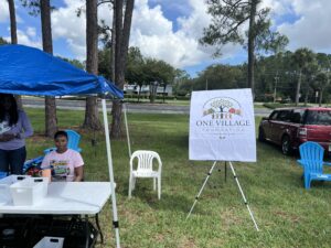 Two women sit at a table under a blue canopy outside, with a sign for “One Village Foundation” on an easel nearby and several empty chairs on the grass Trees and cars are visible in the background