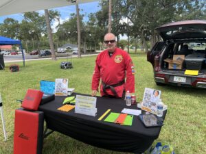 A man in a red martial arts uniform stands behind a table with pamphlets and mats at an outdoor event, with trees and cars visible in the background