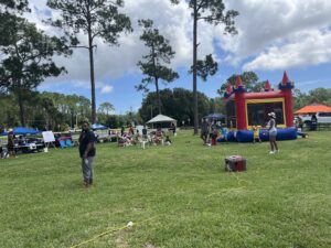 People gather at an outdoor event in a grassy field with a bounce house, tents, and tables Children play while adults stand and sit Tall pine trees and cloudy skies are in the background