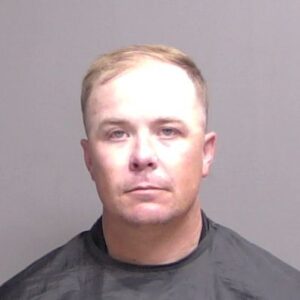 A man with short light hair and a serious expression poses for a mugshot against a plain gray background, wearing a black garment A man with short light hair and a serious expression poses for a mugshot against a plain gray background, wearing a black garment