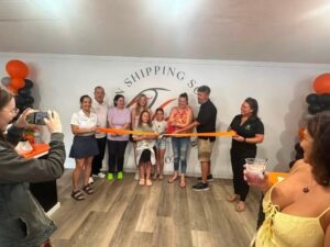 A group of people pose indoors during a ribbon cutting ceremony Some hold the ribbon while others take photos Black and orange balloons decorate the space and a wall sign is partly visible in the background A group of people pose indoors during a ribbon cutting ceremony Some hold the ribbon while others take photos Black and orange balloons decorate the space and a wall sign is partly visible in the background