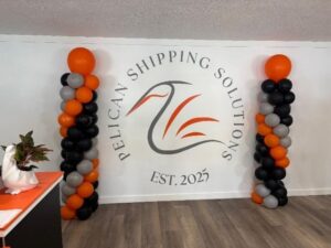 Wall with “Pelican Shipping Solutions Est ” logo featuring a pelican graphic Two balloon columns in orange, black, and gray stand on either side A desk with a white pelican statue is partially visible on the left