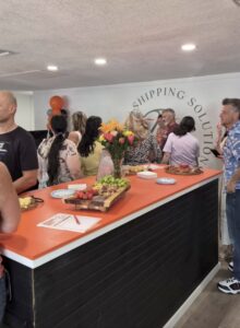 People gather around an orange topped counter with snacks and flowers at a business event Plates and food are on the counter, and a wall says “Shipping Solution” in the background