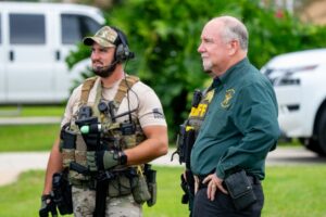 Two law enforcement officers stand outside, one in tactical gear and one in a green sheriff’s uniform, both holding equipment, with vehicles and greenery in the background Two law enforcement officers stand outside, one in tactical gear and one in a green sheriff’s uniform, both holding equipment, with vehicles and greenery in the background