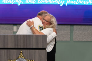 A man in a sheriff’s uniform hugs a smiling woman at a podium A blue screen in the background displays partially visible text