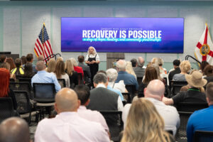 A woman speaks to an audience in a conference room Behind her, a large screen displays “Recovery Is Possible” next to American and Florida flags The audience sits facing her, listening attentively