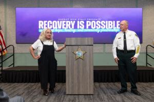 A woman speaks at a podium with a sheriff standing nearby The screen behind them reads, “RECOVERY IS POSSIBLE” and mentions hope, healing, and community partnerships An American flag is visible on the left