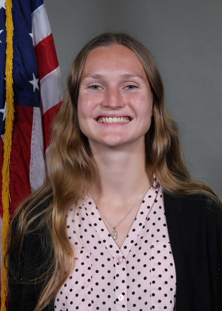 A young woman with long, wavy blonde hair smiles in front of an American flag She wears a light pink blouse with black polka dots and a black cardigan A young woman with long, wavy blonde hair smiles in front of an American flag She wears a light pink blouse with black polka dots and a black cardigan