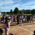 A group of people, some in pajamas, stand and walk outdoors on a sunny day near a fountain, with trees and a building in the background