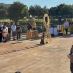 A group of people stand outdoors near a large yellow ribbon sign that reads “Childhood Cancer Awareness ” Some are wearing themed shirts, and a woman is speaking at a table covered with a gold cloth