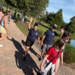 A group of people walk on a park path near a pond Two people in matching navy shirts wave at the camera Others walk ahead, and the sun casts long shadows on the ground Trees and grass surround the area