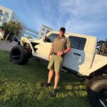 A man in sunglasses, a hat, and green shorts stands smiling by a white off road truck on a grassy area, with modern buildings and a blue sky in the background