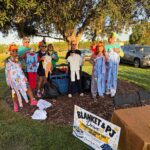 Six people in pajamas and fun hats hold up pajamas at an outdoor Blanket & PJ Drive They stand by a table and a sign under a large tree, with cars and green grass in the background