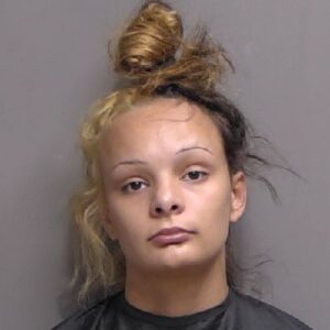 A young woman with a neutral expression, wearing a dark top, has her hair tied up in a messy bun with blonde and dark brown sections, standing against a plain background